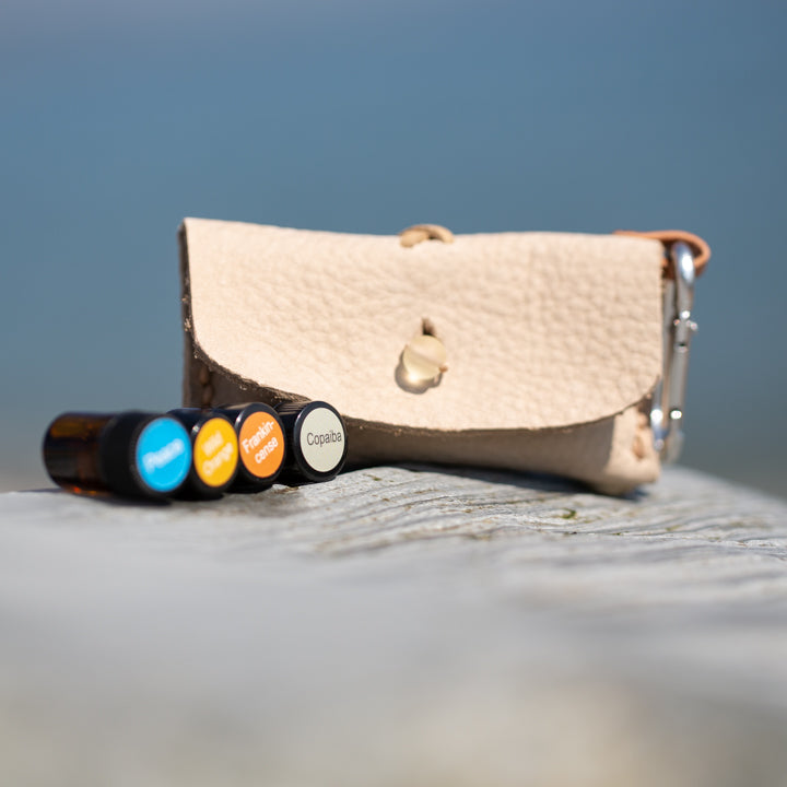 Nature's Essential Gift Kits, small white leather pouch with four 100% natural oils, on a log by the beach.